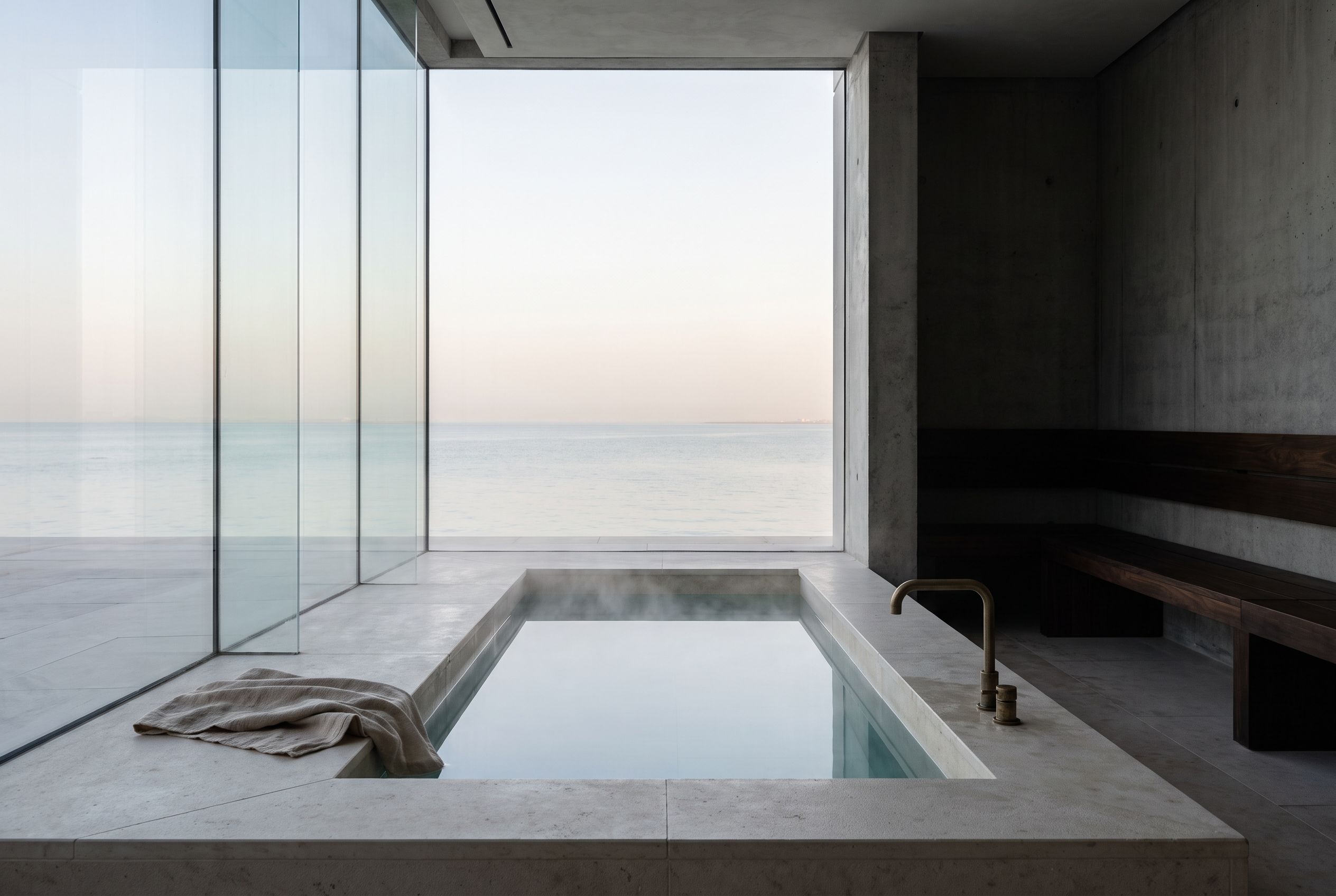 A limestone cold-plunge pool, steam rising, the calm sea beyond the glass at dawn.
